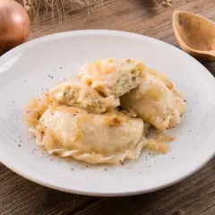 Pierogi z mięsem i cebulką na białym talerzu, obok cebule i drewniane sztućce.