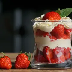Deser z truskawkami i bitą śmietaną w szklance, obok leżą świeże truskawki.