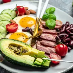Talerz z jajkami sadzonymi, boczkiem, awokado, pomidorkami, ogórkiem i fasolą.