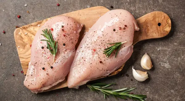Dwie surowe piersi z kurczaka na desce, przyprawione solą, pieprzem i rozmarynem, obok czosnku.