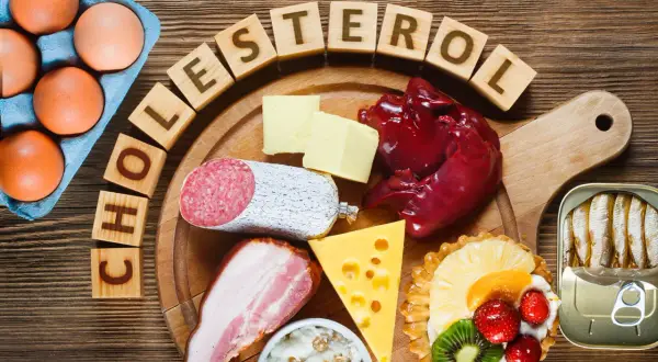 Drewniane klocki z napisem "CHOLESTEROL" otaczają jajka, masło, wątróbkę, kiełbasę, boczek, ser, sardynki i ciastko z owocami.