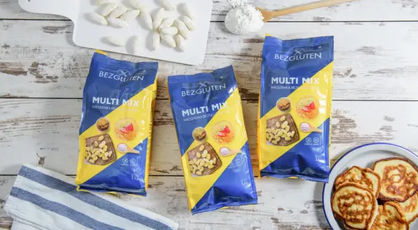 Trzy opakowania mieszanki bezglutenowej Multi Mix, placuszki, mąka i białe paluszki na desce.
