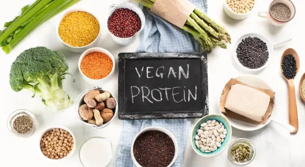 Tablica z napisem "VEGAN PROTEIN" otoczona miseczkami z roślinami strączkowymi, orzechami i warzywami.