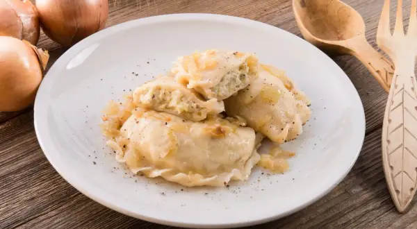 Pierogi z mięsem i cebulką na białym talerzu, obok cebule i drewniane sztućce.