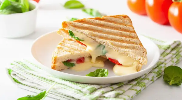 Grillowany tost z serem, pomidorami i szpinakiem na białym talerzu, obok pomidory i miseczka szpinaku.