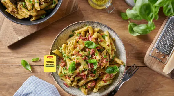 Makaron penne z pesto, boczkiem i bazylią na talerzu, obok słoiczek z pesto i tarką.