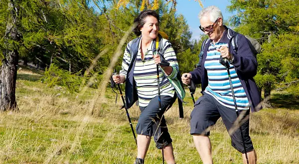 Starsza para wesoło wędruje po lesie z kijkami trekkingowymi.