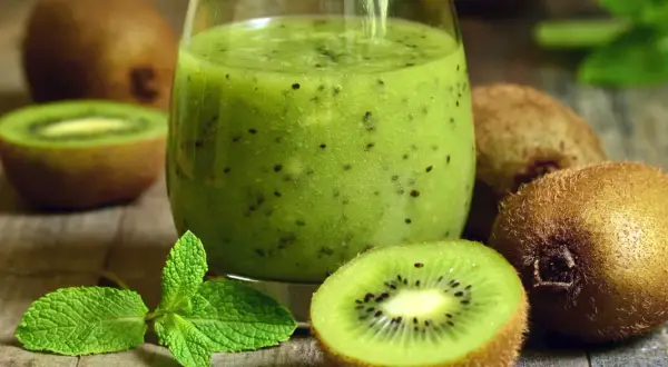 Szklanka zielonego smoothie z kiwi, połówki kiwi i listki mięty na drewnianym tle.
